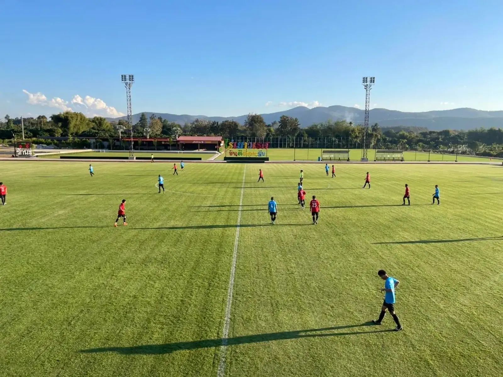 Players training across a full-size grass pitch, floodlight towers and hills behind