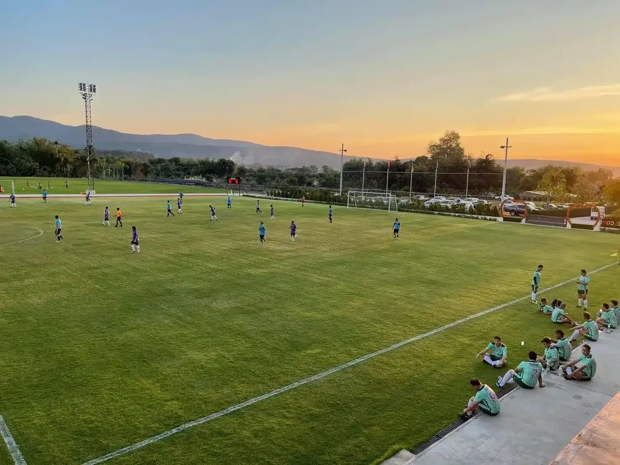 A football squad training on a grass pitch at sunset, with the hills of Mae Taeng behind