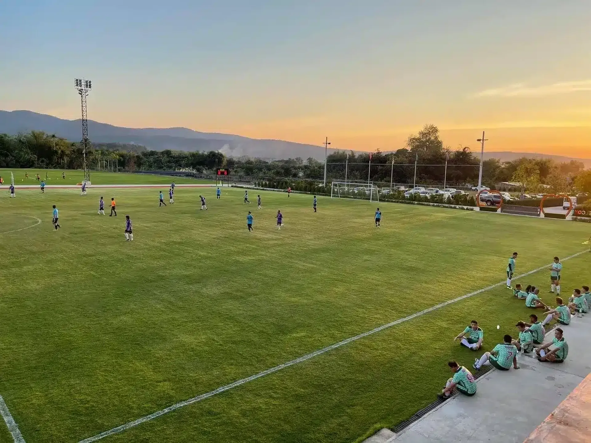 A football squad training on a grass pitch at sunset, with the hills of Mae Taeng behind
